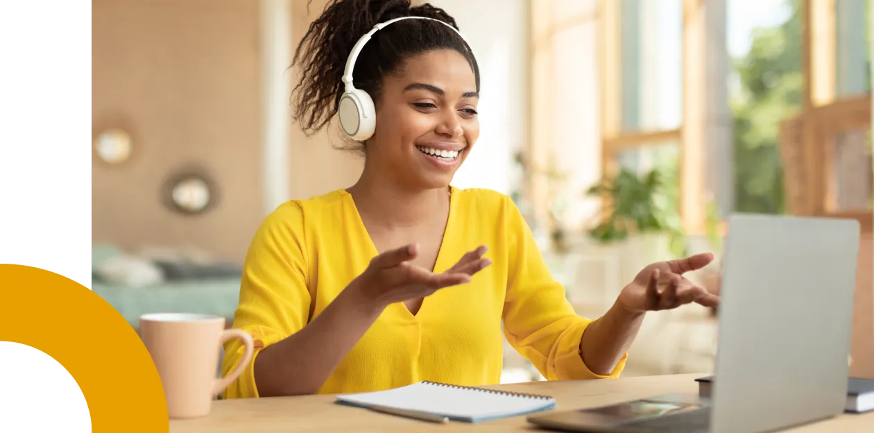 Mujer negra sonriendo en una reunión online con auriculares sentada frente a un escritorio.