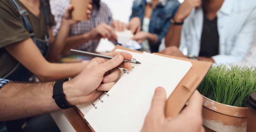 pessoa escrevendo em um caderno com um grupo de pessoas ao fundo