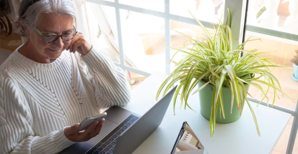 mulher sentada em frente a um notebook com um celular na mão 