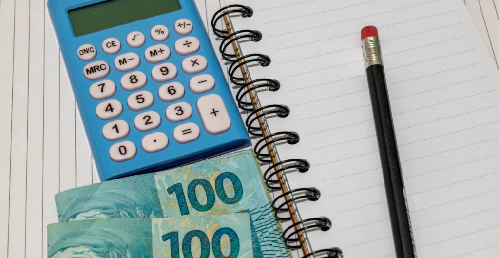 calculadora azul, notas de cem reais e lápis sobre caderno 